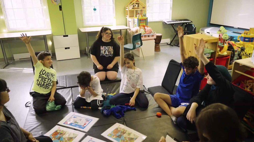 Un jury d'enfants qui votent durant un des ateliers « La force du vote ». Les enfants sont assis sur un tapis, certains ont le bras levé, d'autres pas, tous ont les yeux fermés. 